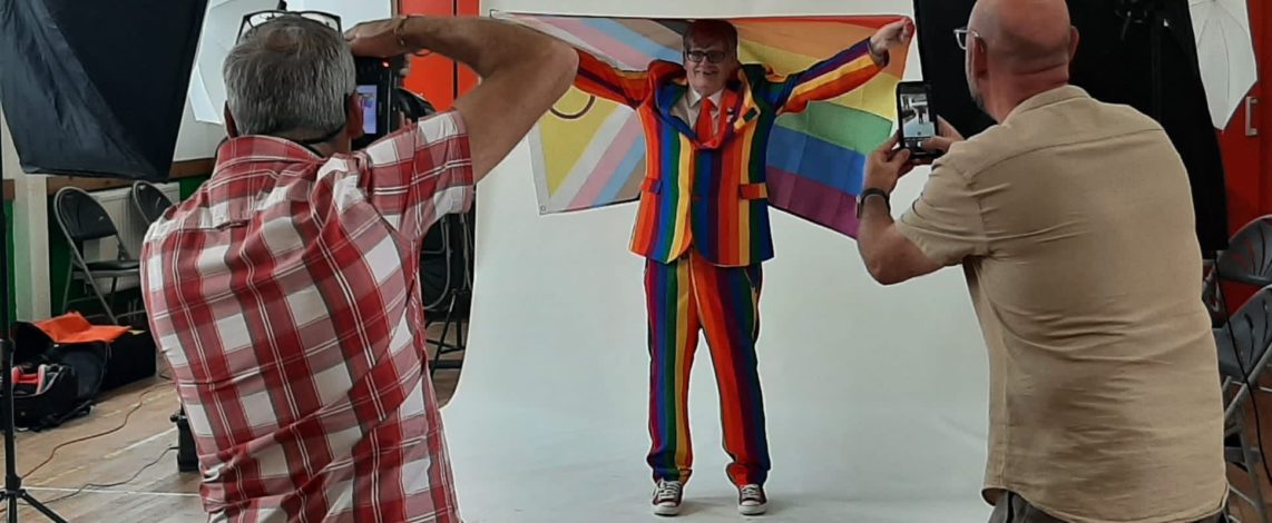 Man in Pride suit holding an inclusive flag with 2 people taking photos of him