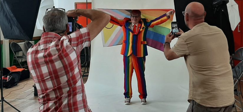 Man in Pride suit holding an inclusive flag with 2 people taking photos of him
