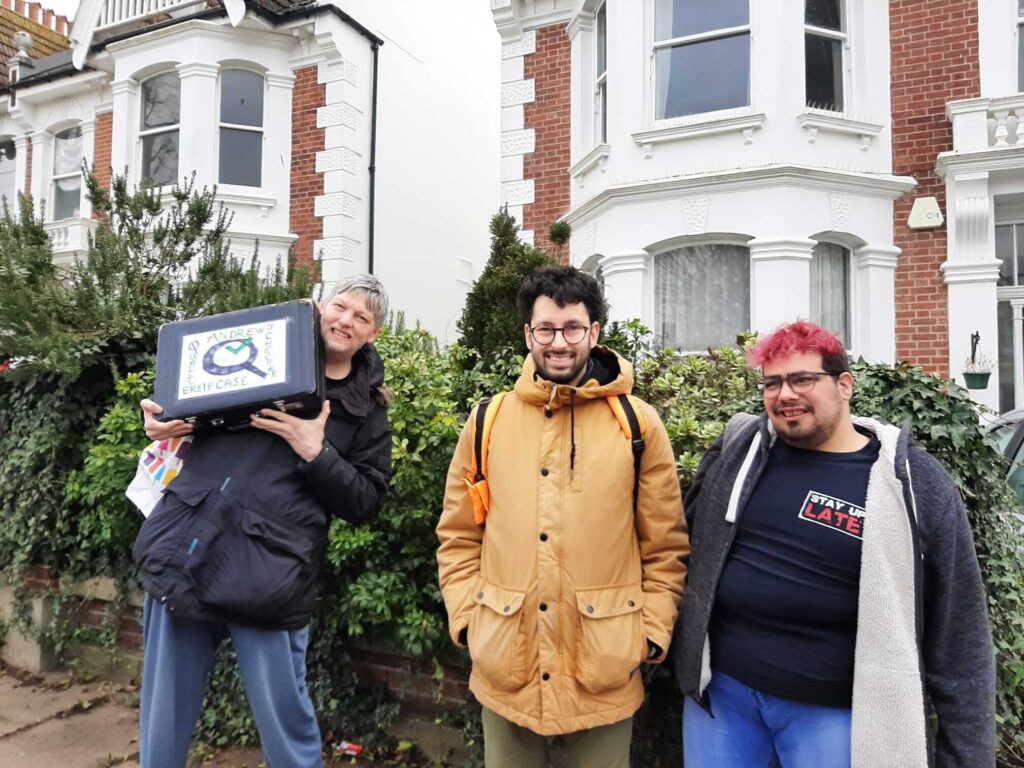 A group of three men stood outside a house. One has a briefcase in his hands that says Quality Checkers on it. Another is wearing a t-shirt that says "Stay Up Late".